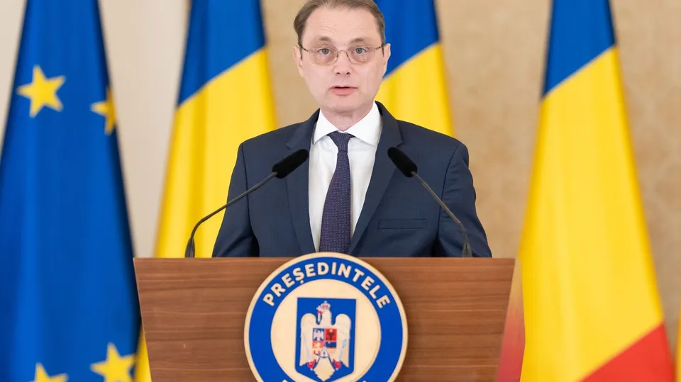 Un bărbat cu ochelari, purtând costum închis și cravată, stă în spatele unui podium de lemn în fața unor steaguri.