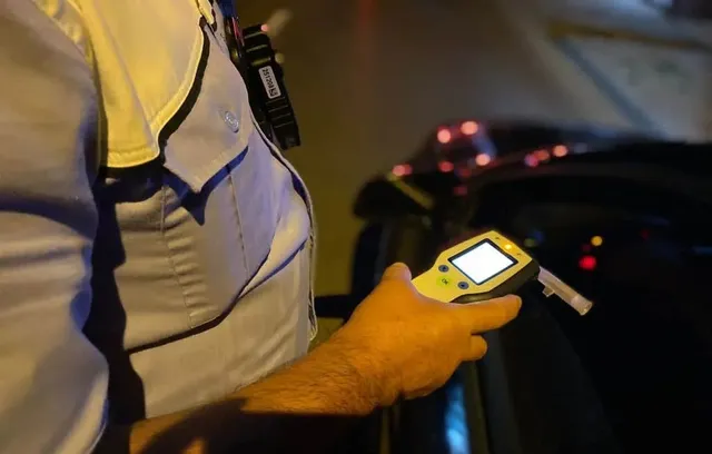 O persoană în uniformă ține un aparat de testare alcolică în mână, în timp ce un vehicul este vizibil în fundalul întunecat.