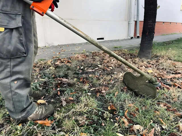 O persoană în pantaloni gri și încălțăminte maro folosește o mașină de tăiat iarba pe un teren acoperit de frunze uscate și iarbă.