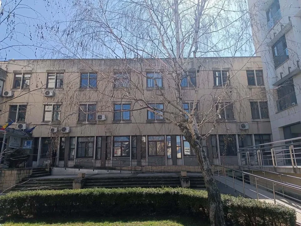 O clădire cu ferestre multiple este parțial acoperită de ramurile unui copac fără frunze în prim-plan.