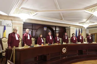 Nouă persoane îmbrăcate în robe roșii și albe stau în spatele unei mese lungi de lemn într-o încăpere cu tavan decorat și corpuri de iluminat.