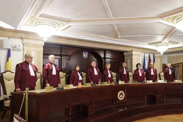 Nouă persoane îmbrăcate în robe roșii și albe stau în spatele unei mese lungi de lemn într-o încăpere cu tavan decorat și corpuri de iluminat.