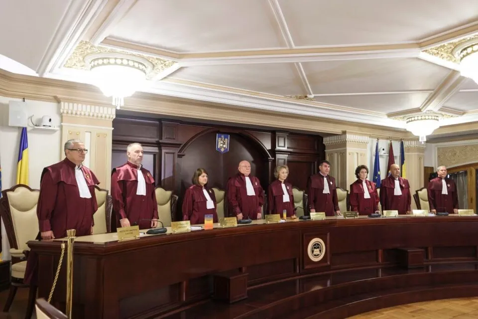 Nouă persoane îmbrăcate în robe roșii și albe stau în spatele unei mese lungi de lemn într-o încăpere cu tavan decorat și corpuri de iluminat.