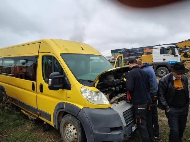 O autoutilitară galbenă cu capota deschisă este verificată de mai bărbați într-un spațiu deschut, cu alte vehicule în fundal.