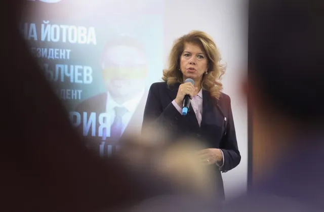 O femeie în costum închis vorbește într-un microfon, pe fundal fiind un afiș cu text și o imagine a unui bărbat.