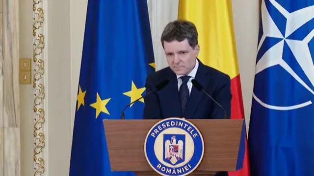Un bărbat cu părul scurt și costum închis la culoare stă în spatele unui podium cu emblema prezidențială, în fața unor steaguri.