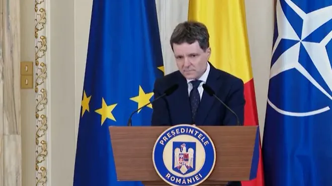 Un bărbat cu părul scurt și costum închis la culoare stă în spatele unui podium cu emblema prezidențială, în fața unor steaguri.