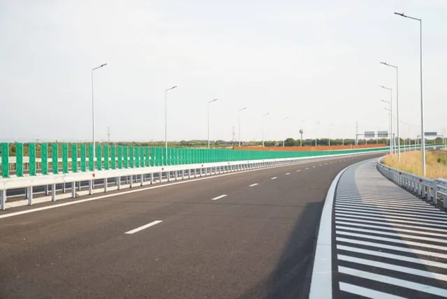 O autostradă cu asfalt negru, linii albe marcate și parapeti metalici curbează spre dreapta sub un cer deschis.