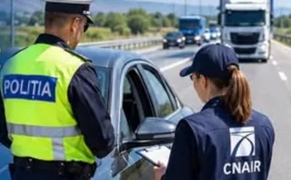 Un polițist cu vestă reflectorizantă și o femeie cu șapcă și vestă CNAIR stau lângă o mașină gri pe un drum cu trafic.