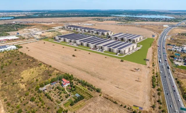 O vedere aeriană prezintă un complex de clădiri industriale cu panouri solare pe acoperiș, situat lângă o autostradă și un câmp gol.
