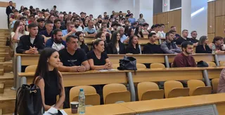 Un grup de persoane este așezat pe scaune în interiorul unei săli de curs cu birouri de lemn dispuse în trepte.