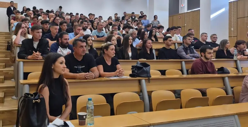 Un grup de persoane este așezat pe scaune în interiorul unei săli de curs cu birouri de lemn dispuse în trepte.