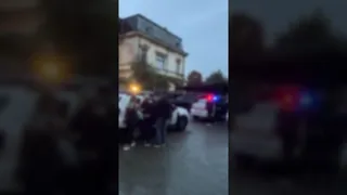 Un grup de persoane stă lângă mașini cu lumini de alertă aprinse în fața unei clădiri sub un cer închis.