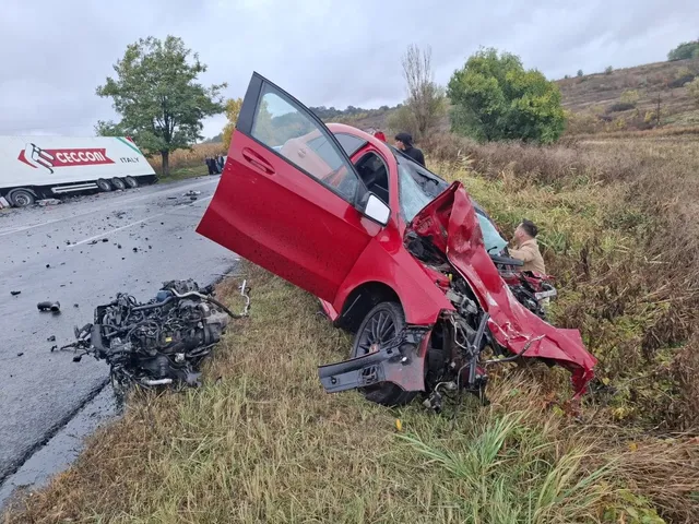 Un autoturism roșu avariat stă pe marginea unui drum, cu portiera deschisă și motorul separat pe iarbă, lângă un camion alb.