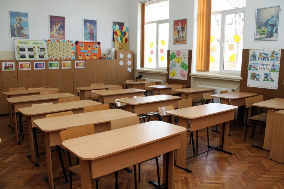 O sală de clasă cu bănci de lemn dispuse în rânduri și diverse afișe colorate afișate pe pereți și pe un panou de lemn.