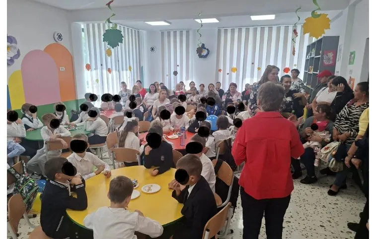 Un grup de copii și adulți stau la mese rotunde într-o încăpere iluminată de ferestre mari cu jaluzele verticale albe.