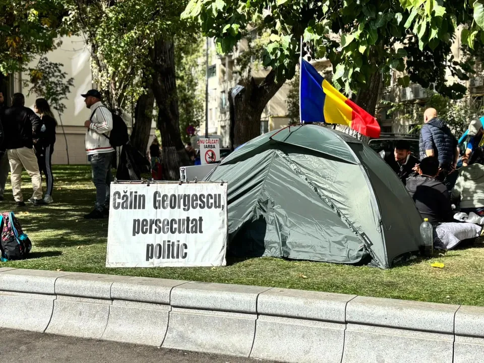 Un cort gri este amplasat pe iarbă lângă un banner alb cu text negru, sub copaci, lângă un steag tricolor.