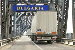 Un camion alb cu remorcă circulă pe un pod metalic de tip pod de oțel lângă un panou albastru pe care scrie text galben.