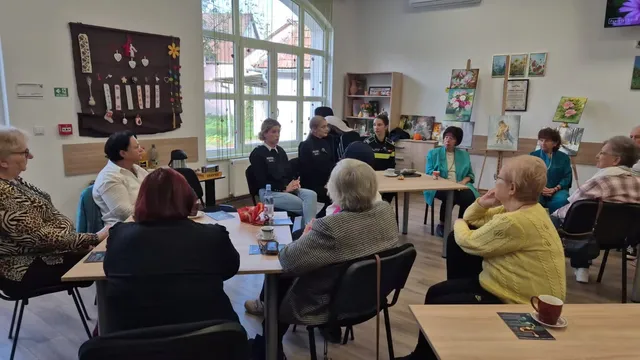 Un grup de persoane stă la mese în interiorul unei încăperi, orientate spre un grup de oameni care se află în partea opusă a camerei.