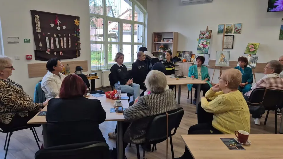 Un grup de persoane stă la mese în interiorul unei încăperi, orientate spre un grup de oameni care se află în partea opusă a camerei.