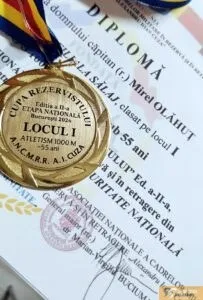 O medalie aurie cu inscripții este așezată pe o diplomă albă cu text imprimat negru și un fundal albastru în partea de sus.