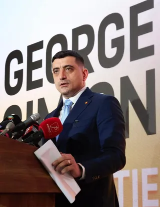 Un bărbat într-un costum albastru și cămașă albastră stă în spatele unui podium cu microfoane și ține o foaie de hârtie albă.