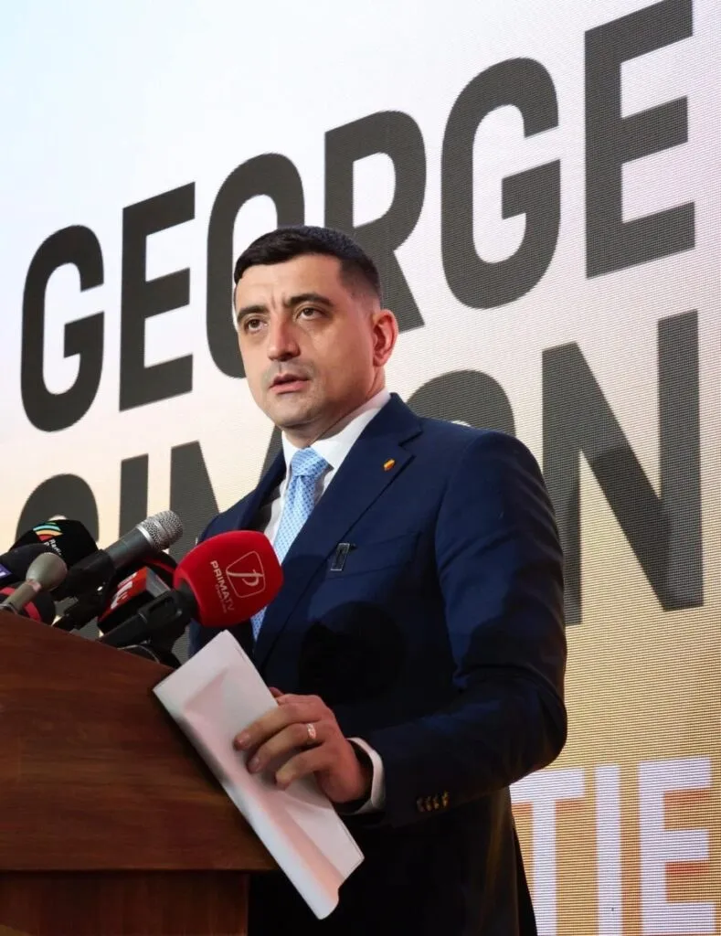 Un bărbat într-un costum albastru și cămașă albastră stă în spatele unui podium cu microfoane și ține o foaie de hârtie albă.