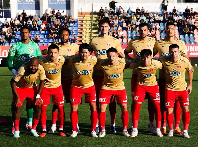 Un grup de fotbaliști în echipament sportiv galben și roșu pozează într-o poziție de echipă pe un teren de fotbal verde.