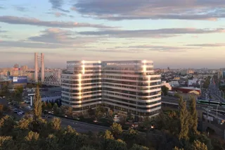 O clădire de birouri modernă cu fațadă de sticlă și linii orizontale albe este situată într-un peisaj urban sub un cer la apus.