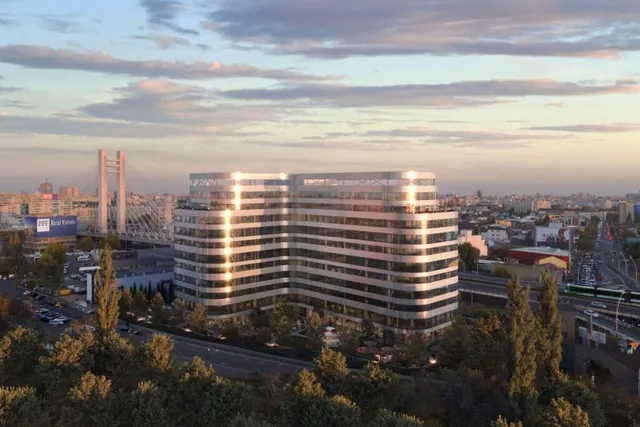 O clădire de birouri modernă cu fațadă de sticlă și linii orizontale albe este situată într-un peisaj urban sub un cer la apus.