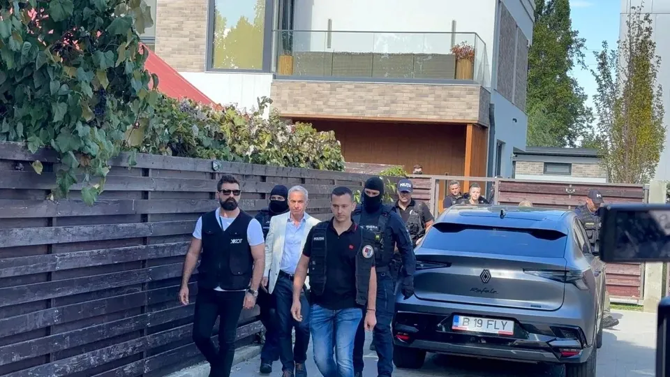 Un grup de bărbați merge pe un drum lângă un gard de lemn și o clădire modernă, lângă o mașină gri în parcarea exterioară.