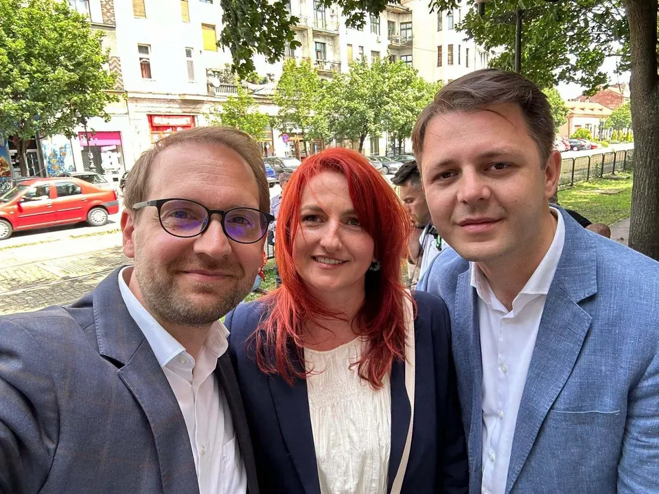 Trei persoane stau apropiate una de alta, un bărbat cu ochelari, o femeie cu părul roșu și un bărbat într-un sacou albastru.
