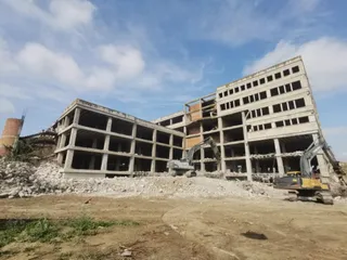 Două excavatoare efectuează lucrări de demolare lângă o clădire de beton cu mai multe etaje sub un cer cu nori.