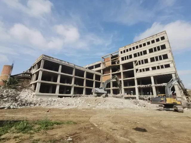 Două excavatoare efectuează lucrări de demolare lângă o clădire de beton cu mai multe etaje sub un cer cu nori.