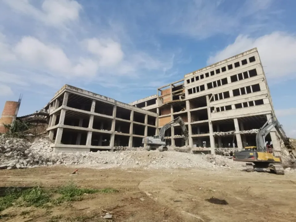 Două excavatoare efectuează lucrări de demolare lângă o clădire de beton cu mai multe etaje sub un cer cu nori.