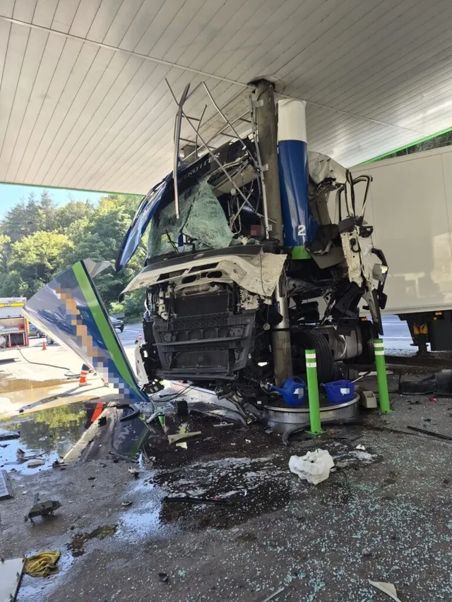 Un camion alb și albastru este zdrobit sub acoperișul unei stații de alimentare, fiind înconjurat de resturi și lichid pe asfaltul umed.