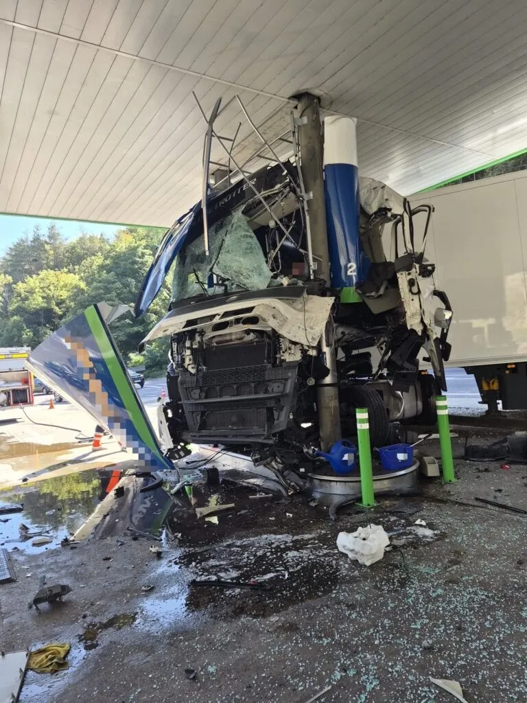Un camion alb și albastru este zdrobit sub acoperișul unei stații de alimentare, fiind înconjurat de resturi și lichid pe asfaltul umed.