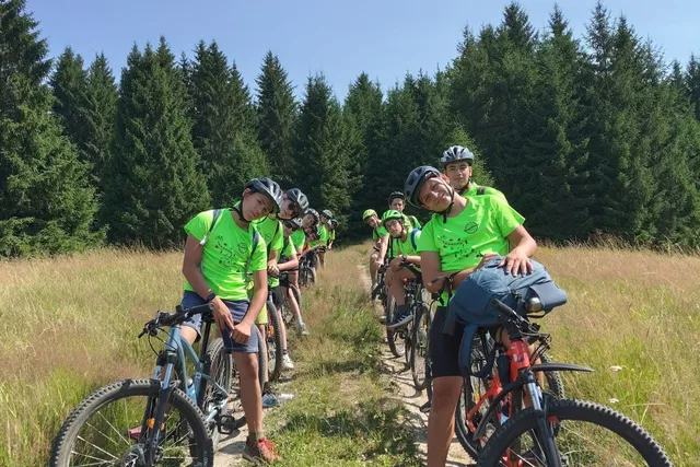 Un grup de tineri cicliști, purtând tricouri verzi și căști, stau pe biciclete pe o potecă îngustă, înconjurați de iarbă înaltă și copaci.