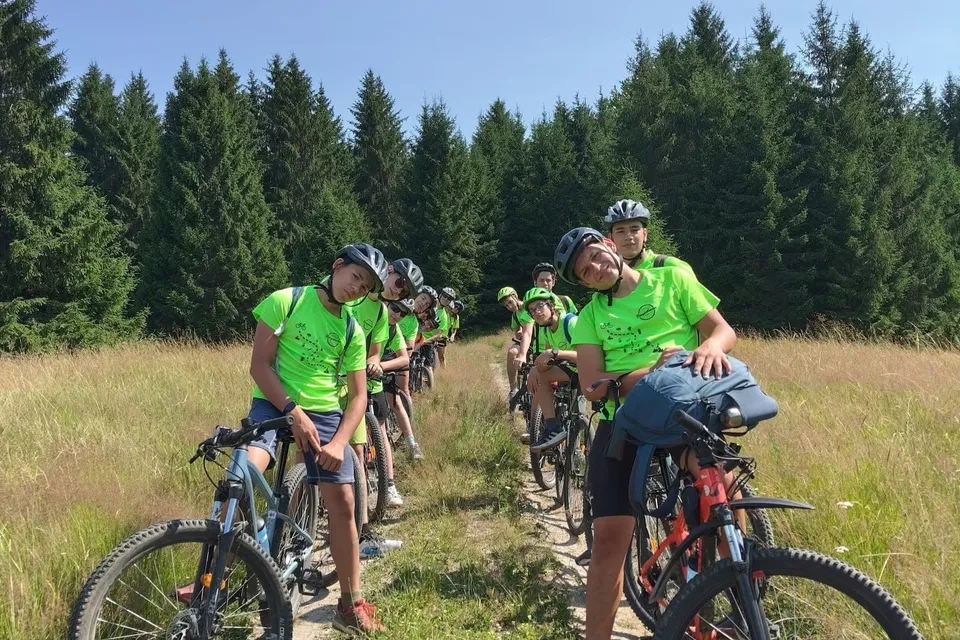 Un grup de tineri cicliști, purtând tricouri verzi și căști, stau pe biciclete pe o potecă îngustă, înconjurați de iarbă înaltă și copaci.