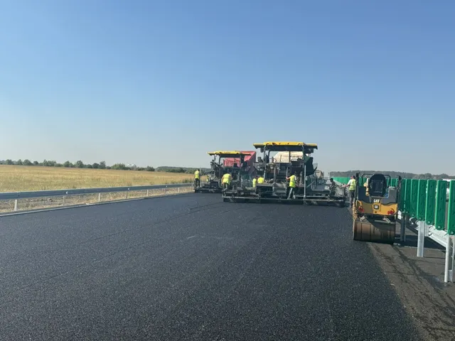 Mașini de asfaltare și un cilindru de compactare lucrează pe o suprafață de asfalt proaspăt turnat sub un cer senin.