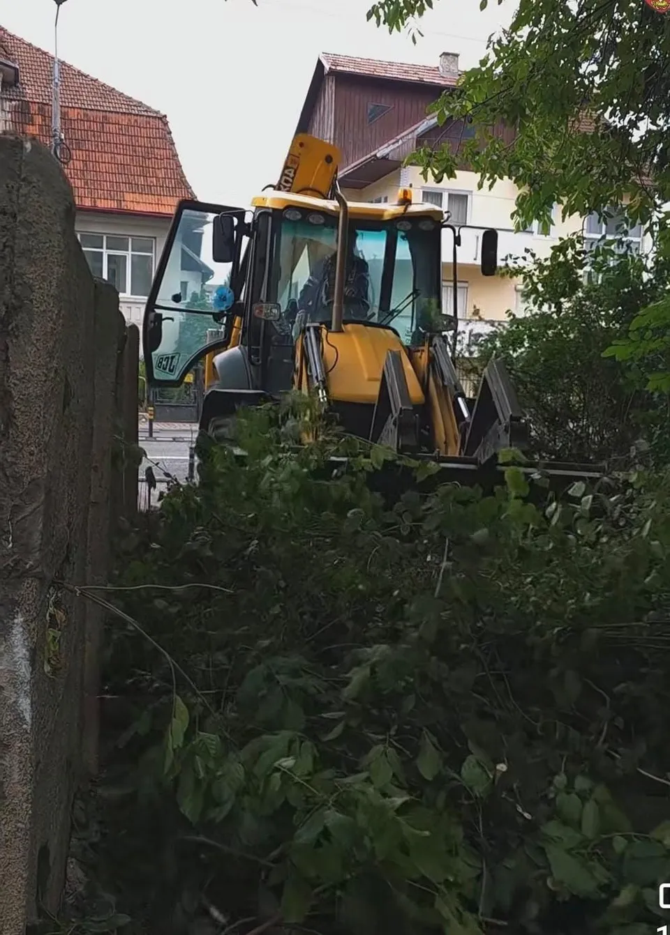 Un excavator galben este oprit lângă un gard, în fața unor arbori și a unor clădiri rezidențiale.