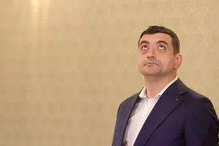 Un bărbat cu părul scurt și sacou albastru privește în sus în fața unui perete cu tapet decorativ.
