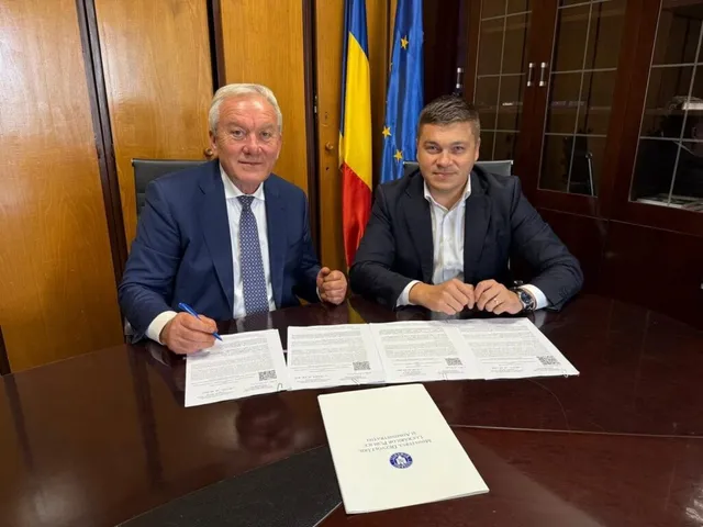 Un bărbat cu părul alb și unul cu părul scurt stau la o masă de lemn lângă două steaguri, având documente și un dosar în față.