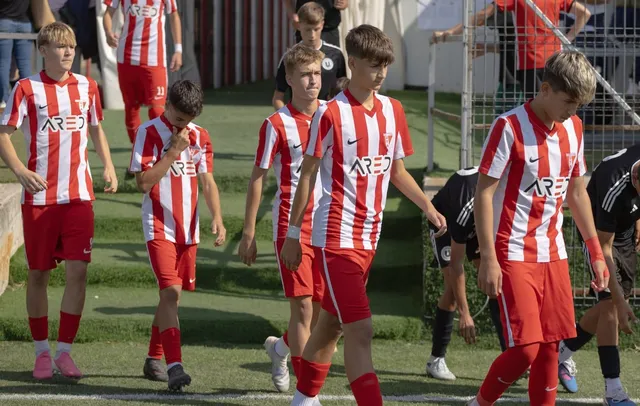 Un grup de tineri sportivi îmbrăcați în echipamente de fotbal cu tricouri cu dungi roșii și albe și pantaloni scurți roșii merg pe un teren.