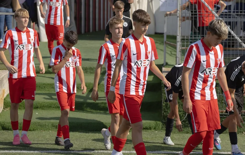 Un grup de tineri sportivi îmbrăcați în echipamente de fotbal cu tricouri cu dungi roșii și albe și pantaloni scurți roșii merg pe un teren.