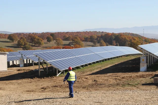 Un lucrător cu vestă reflectorizantă merge pe un teren de pământ lângă un rând de panouri solare instalate pe suporturi metalice.