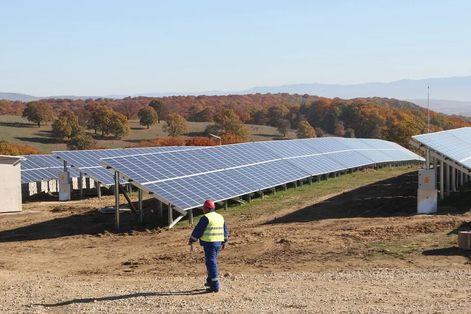 Un lucrător cu vestă reflectorizantă merge pe un teren de pământ lângă un rând de panouri solare instalate pe suporturi metalice.