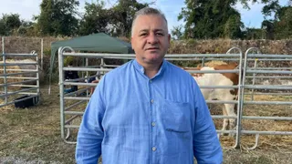 Un bărbat cu cămașă albastră stă în fața unor țarcuri metalice care conțin animale într-un câmp cu vegetație și copaci în fundal.