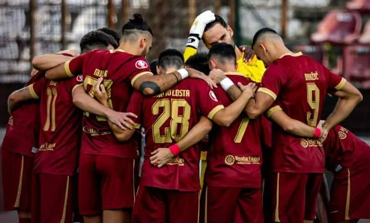 O echipă de fotbaliști în echipamente de culoare roșu închis stau într-un cerc, cu brațele pe umerii colegilor.
