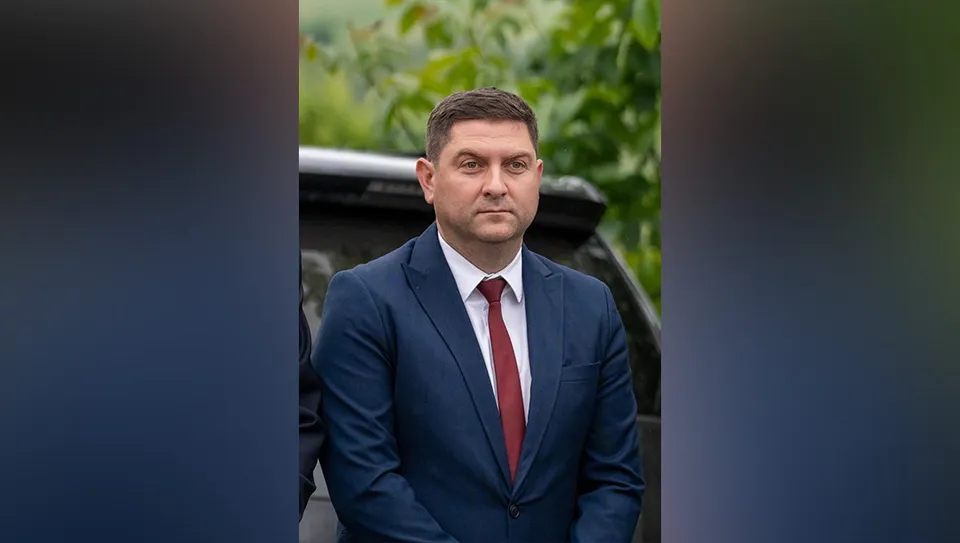 Un bărbat cu părul scurt, purtând un costum albastru, cămașă albă și cravată roșie, stă în fața unui vehicul negru.